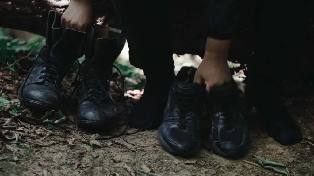 Close up of the feet of a person sitting on a log in the forest with a pair of old fashioned black boots in one hand and a pair of modern black trainers in the other.